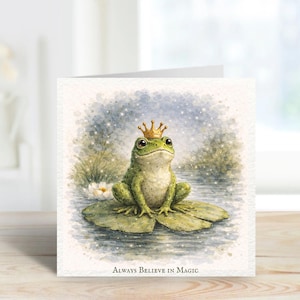 Puede incluir: Tarjeta de felicitación con una ilustración en acuarela de una rana verde con una corona dorada, sentada en una hoja de nenúfar. El texto "Always Believe in Magic" está en la parte inferior.