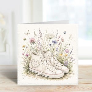 May include: A greeting card featuring a watercolor illustration of white high-top sneakers with white laces. The sneakers are surrounded by wildflowers, grasses, and butterflies. The text "Just Married" is printed on the side of one sneaker.