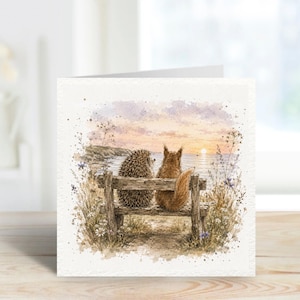 May include: A watercolour greeting card depicting a hedgehog and a squirrel seated on a wooden bench, observing a sunset over the sea. The image features wildflowers and a soft, pastel colour scheme.