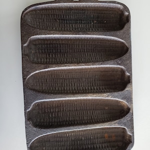 May include: A cast iron cornbread pan with five corn cob molds. The pan is dark brown and has a handle with a hole for hanging. The molds are detailed with the texture of corn kernels.