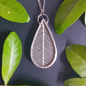Könnte beinhalten: Eine silberne Kettenkette mit einem Anhänger, der ein zartes, getrocknetes Blatt in klarem Harz enthält. Das Blatt hat ein detailliertes Adernmuster und ist tropfenförmig.