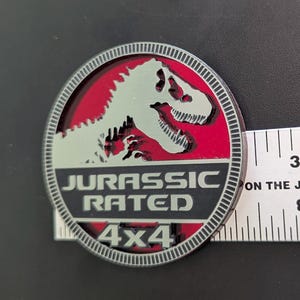 May include: A round, silver-edged emblem with a red center featuring a dinosaur silhouette. The text "JURASSIC RATED 4X4" is displayed in white. The emblem is approximately 3 inches in diameter.