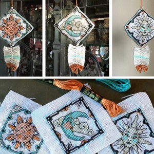 Puede incluir: Tres tapices de pared bordados con un diseño de sol, luna y nube. Cada tapiz tiene un diseño de plumas azul y blanco en la parte inferior. Los tapices están sobre un fondo blanco.