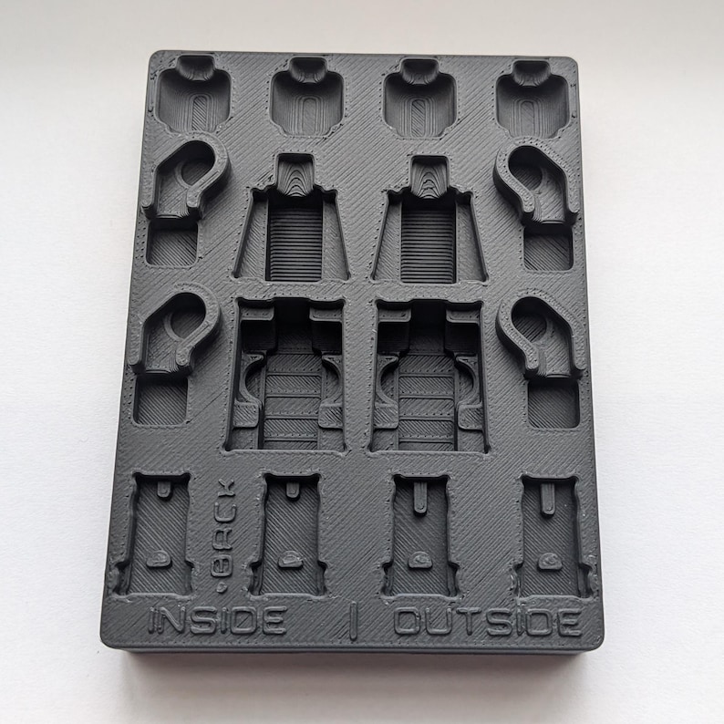 May include: A black plastic mold with multiple cavities for creating small figures. The mold has sections labeled "INSIDE" and "OUTSIDE" with the word "BACK" above. The cavities are shaped like miniature torsos, arms, and heads.