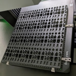 May include: A dark gray plastic tray filled with small, cutout figures, possibly for a manufacturing process. The tray is held in place with white clips and has measurement markings along the edge. The text "MAX kg" is visible.