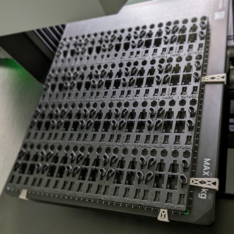 May include: A dark gray plastic tray filled with small, cutout figures, possibly for a manufacturing process. The tray is held in place with white clips and has measurement markings along the edge. The text "MAX kg" is visible.