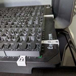 May include: A close-up of a black, rectangular object with a grid-like pattern, possibly a keyboard component. White clips secure the object to a black base with a measurement scale. The text "MAX kg" is visible.