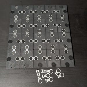 May include: A dark gray interlocking puzzle board with a grid pattern of connecting pieces. The board has a textured surface and is partially assembled. Several white connecting pieces are scattered nearby.