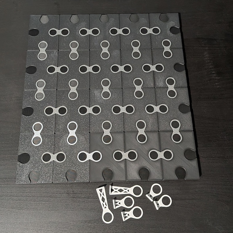 May include: A dark gray interlocking puzzle board with a grid pattern of connecting pieces. The board has a textured surface and is partially assembled. Several white connecting pieces are scattered nearby.