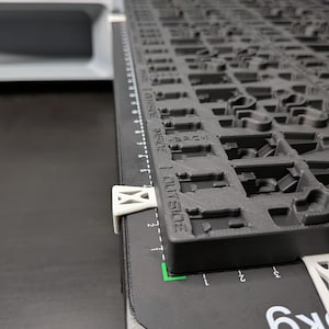 May include: A black and gray 3D-printed keyboard layout with a ruler along the side. The keyboard layout has various cutouts for key placement. White clips secure the keyboard layout to a black surface. The ruler is marked in inches.