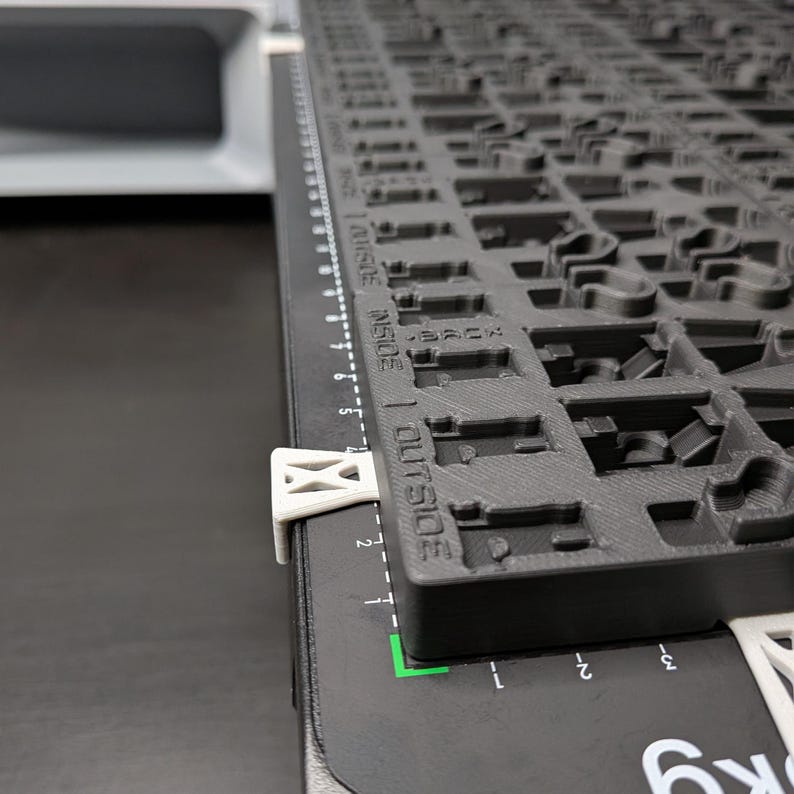 May include: A black and gray 3D-printed keyboard layout with a ruler along the side. The keyboard layout has various cutouts for key placement. White clips secure the keyboard layout to a black surface. The ruler is marked in inches.