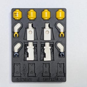 May include: A black plastic organizer tray holding disassembled yellow Lego heads, white torsos, and white arms with yellow hands. The tray is labeled "INSIDE" and "OUTSIDE".