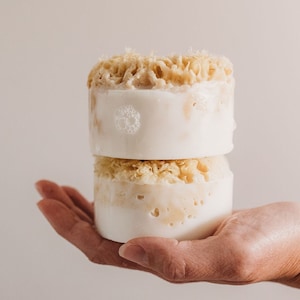 May include: Two white and beige round soap bars stacked on top of each other. The top bar has a beige sponge-like texture. The bottom bar has a similar texture but is slightly more compressed. The bars are held in a hand.