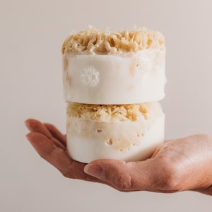 May include: Two white and beige soap bars stacked on top of each other. The top bar has a beige sponge-like texture. The bottom bar has a similar texture but is slightly more compressed. The bars are held in a hand.
