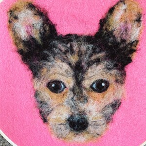 May include: A needle felted portrait of a dog with brown and black fur, on a pink background.