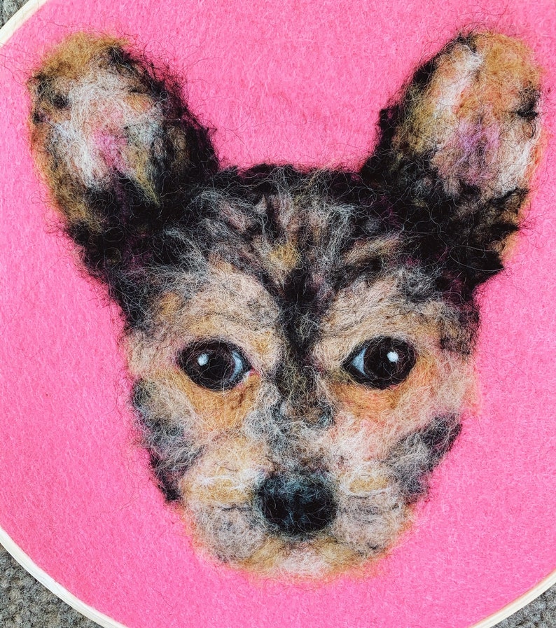 May include: A needle felted portrait of a dog with brown and black fur, on a pink background.