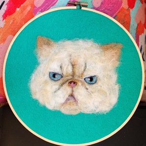 May include: A white, fluffy cat with blue eyes is depicted in a needle felted portrait. The cat has a grumpy expression and is framed in a wooden embroidery hoop with a turquoise background.