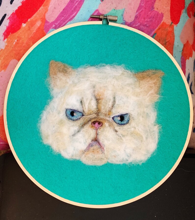 May include: A white, fluffy cat with blue eyes is depicted in a needle felted portrait. The cat has a grumpy expression and is framed in a wooden embroidery hoop with a turquoise background.