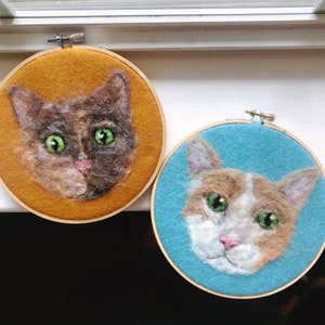 May include: Two needle felted cat portraits in embroidery hoops. The cat on the left is brown and black with green eyes and is on a yellow background. The cat on the right is orange and white with green eyes and is on a blue background.