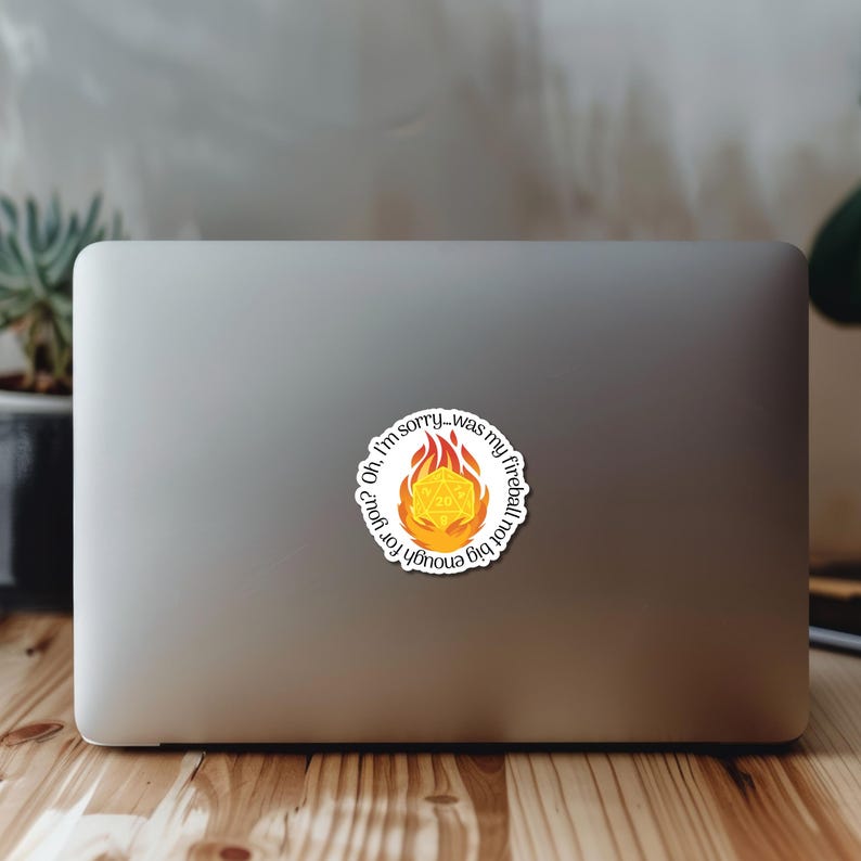 May include: A white sticker on a laptop. The sticker features a yellow and orange fireball design with a D20 die in the center. The text "Oh, I'm sorry...was my fireball not big enough for you?" is in a circular pattern.