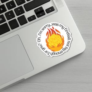 May include: A white sticker with the text "Oh, I'm sorry...was my fireball not big enough for you?" surrounds a yellow 20-sided die engulfed in flames. The sticker is on a silver laptop.