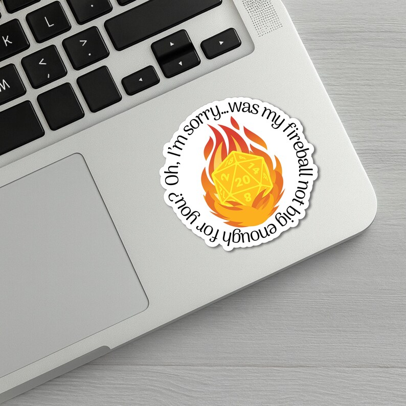 May include: A white sticker with the text "Oh, I'm sorry...was my fireball not big enough for you?" surrounds a yellow 20-sided die engulfed in flames. The sticker is on a silver laptop.