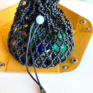 May include: Black beaded dice bag with a drawstring closure and metal tips. The bag is filled with colorful dice and is sitting on a yellow felt tray.