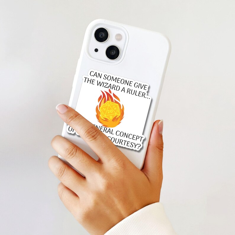 May include: White phone case with a sticker featuring a yellow 20-sided die engulfed in flames. The sticker has the text "CAN SOMEONE GIVE THE WIZARD A RULER..." and "GENERAL CONCEPT COURTESY?"