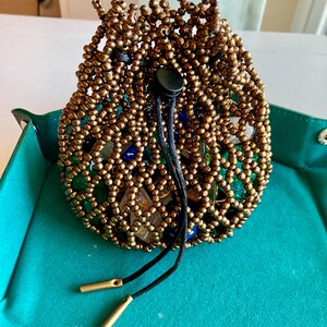 May include: A beaded drawstring pouch in a bronze color, filled with dice. The pouch is made of small, round beads, and the drawstring is black with gold-colored tips. The pouch sits on a teal surface.