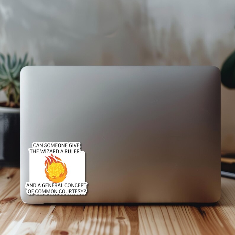 May include: A sticker on a laptop features a graphic of a flaming, yellow, multi-sided die. Text on the sticker reads, "CAN SOMEONE GIVE THE WIZARD A RULER... AND A GENERAL CONCEPT OF COMMON COURTESY?" The sticker is white with black text.