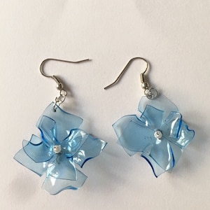 May include: A pair of blue flower-shaped earrings made from recycled plastic. The earrings have silver hooks and a small white bead in the centre of each flower.