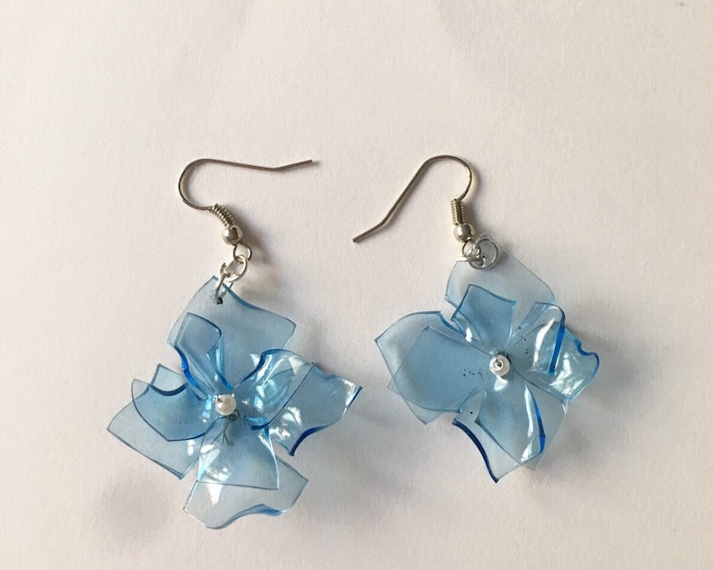 May include: A pair of blue flower-shaped earrings made from recycled plastic. The earrings have silver hooks and a small white bead in the centre of each flower.