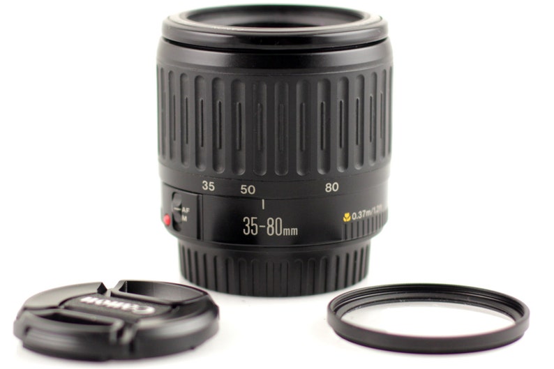 May include: A black Canon EF 35-80mm f/4-5.6 lens with a black lens cap and a black filter ring. The lens has a zoom ring and a focus ring. The lens is designed for Canon EOS cameras.