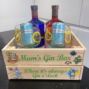 Puede incluir: Una caja de madera con las palabras "Mum's Gin Bar" y "Where it's always Gin o'clock" impresas en ella. La caja contiene dos vasos de vidrio transparentes, dos botellas de ginebra y dos latas de agua tónica.