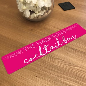 May include: A rectangular, hot pink cocktail bar sign with the text "THE HARRISON'S cocktail bar" in white script. Decorative flourishes are at each end of the sign. The sign is on a wooden surface.