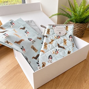 May include: A light blue quilted oven mitt and matching kitchen towel set featuring a repeating pattern of various dog breeds. The oven mitt has a round tag labeled Cooksmart Gauntlet. The items are neatly arranged inside a white rectangular box.