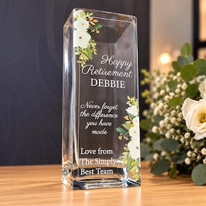 Puede incluir: Un florero rectangular de vidrio transparente con un diseño floral de flores blancas y hojas verdes. El texto dice: Happy Retirement DEBBIE, Never forget the difference you have made, Love from The Simply Best Team, impreso en letras blancas.
