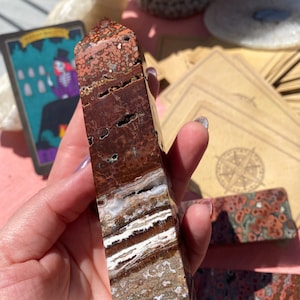 May include: A polished, rectangular gemstone tower with a mix of red, brown, white, and tan colors. The stone has a layered pattern with unique textures and small holes. The tower is held in a hand, with a tarot card and other stones in the background.