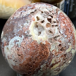 May include: A large, round, brown, white, and pink agate sphere with a large, open, white crystal geode in the center. The sphere is sitting on a black stand.