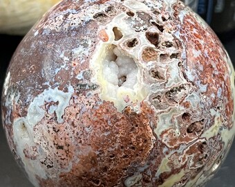 May include: A large, round, brown, white and pink agate sphere with a large, open, white crystal geode in the centre. The sphere is sitting on a black stand.