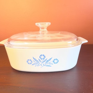 Op de afbeelding: Een witte rechthoekige glazen ovenschaal met geïntegreerde handgrepen aan de zijkant en een doorzichtig glazen deksel met een knop. De voorzijde van de schaal is versierd met een blauw bloemmotief van drie gestileerde bloemen en bladeren.