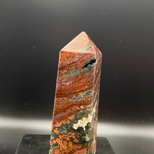 Könnte beinhalten: Ein polierter, rechteckiger Edelstein-Obelisk. Der Stein weist eine Mischung aus rotbraunen, grünen und cremefarbenen Farben mit einem geschichteten Muster auf. Die Oberseite hat eine spitze, abgewinkelte Form. Der Obelisk steht aufrecht.