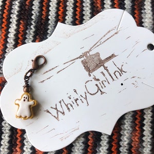 Op de afbeelding: Witte spookvormige hanger met een koperen ketting en sluiting bevestigd aan een wit bord met de tekst "Whirly Girl Ink".