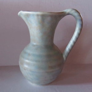 May include: A small, light blue ceramic jug with a textured, swirling design. The jug has a rounded body and a slightly flared lip. The handle is twisted and has a light brown colour.