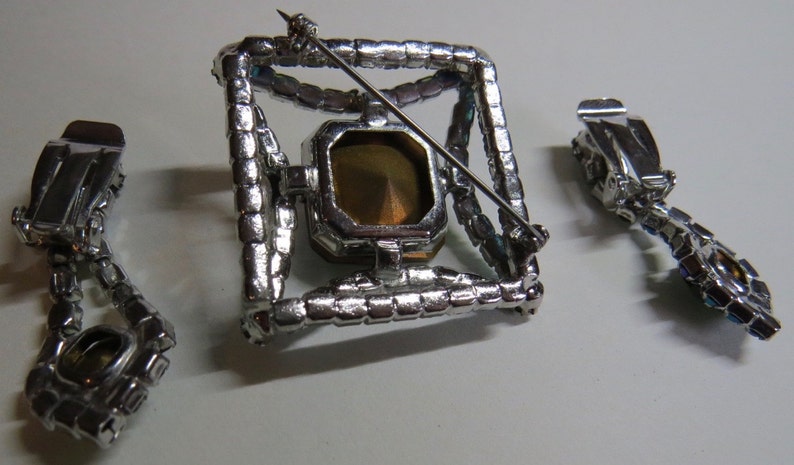 Puede incluir: Un conjunto de broche y pendientes de tono plateado con una piedra grande, transparente y facetada en un marco cuadrado. El broche tiene un cierre de pin y los pendientes tienen cierres de clip.
