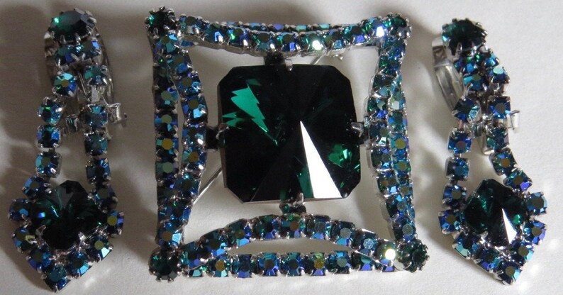 Puede incluir: Un conjunto de broche y pendientes vintage con un gran strass verde esmeralda en un marco cuadrado rodeado de peque&ntilde;os strass azules.