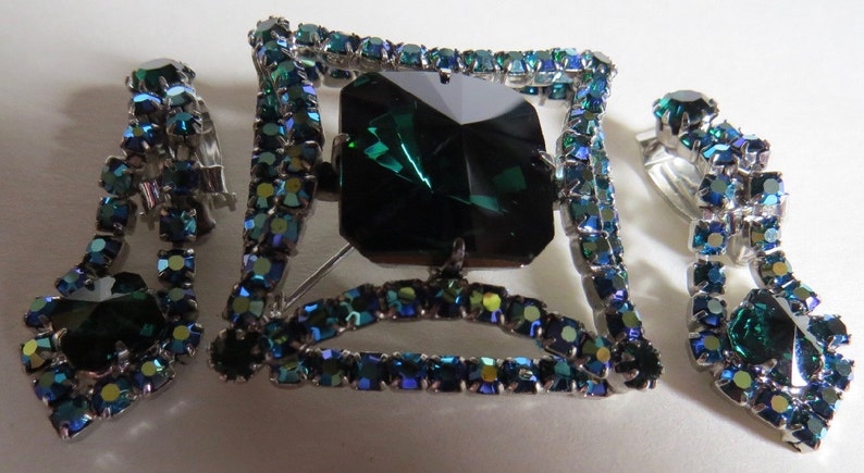 Puede incluir: Un conjunto de joyas de pedrer&iacute;a verde esmeralda y azul con un broche cuadrado con una gran piedra verde esmeralda y pendientes de clip a juego.