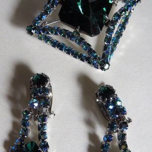Puede incluir: Un conjunto de joyas de pedrer&iacute;a verde esmeralda que incluye un broche cuadrado con una gran pedrer&iacute;a verde esmeralda en el centro y dos pendientes colgantes a juego.