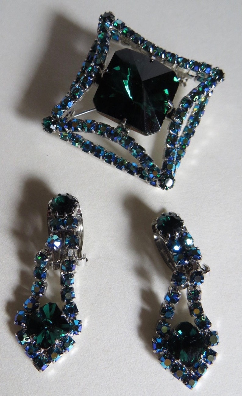 Puede incluir: Un conjunto de joyas de pedrer&iacute;a verde esmeralda que incluye un broche cuadrado con una gran pedrer&iacute;a verde esmeralda en el centro y dos pendientes colgantes a juego.