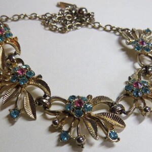 May include: Vintage gold-tone necklace featuring floral clusters with blue and pink rhinestones. The necklace has a delicate chain and is a classic piece of jewelry.